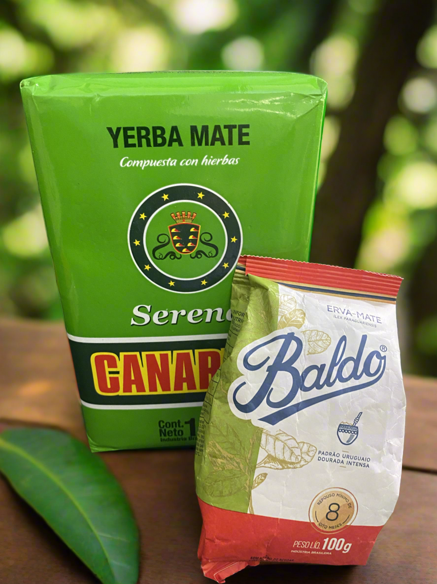 Two packages of yerba mate. One labeled 'Canarias Serena' in green and the other 'Baldo' in white and red, both on a wooden surface with foliage in the background.
