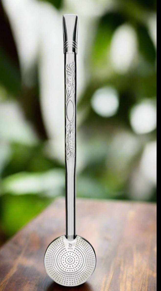 A silver bombilla with 1000 holes designed for straining yerba mate, featuring intricate engravings and a circular filter at the bottom, displayed against a blurred greenery background.