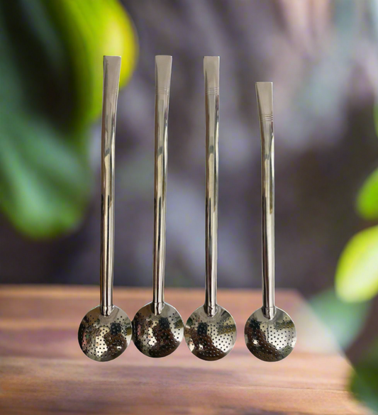 A collection of four stainless steel bombillas with strainer balls, displayed upright on a wooden surface with a blurred green plant background.