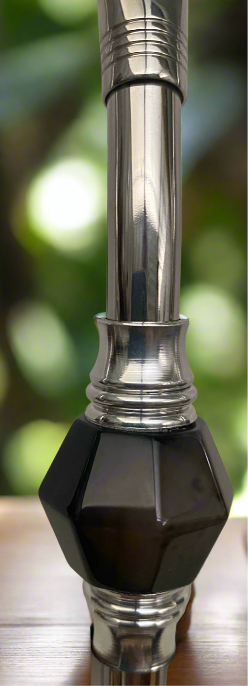 A stainless steel bombilla dark brown in color, featuring a long handle and a dark brown stone filter, placed on a wooden surface with a blurred background.