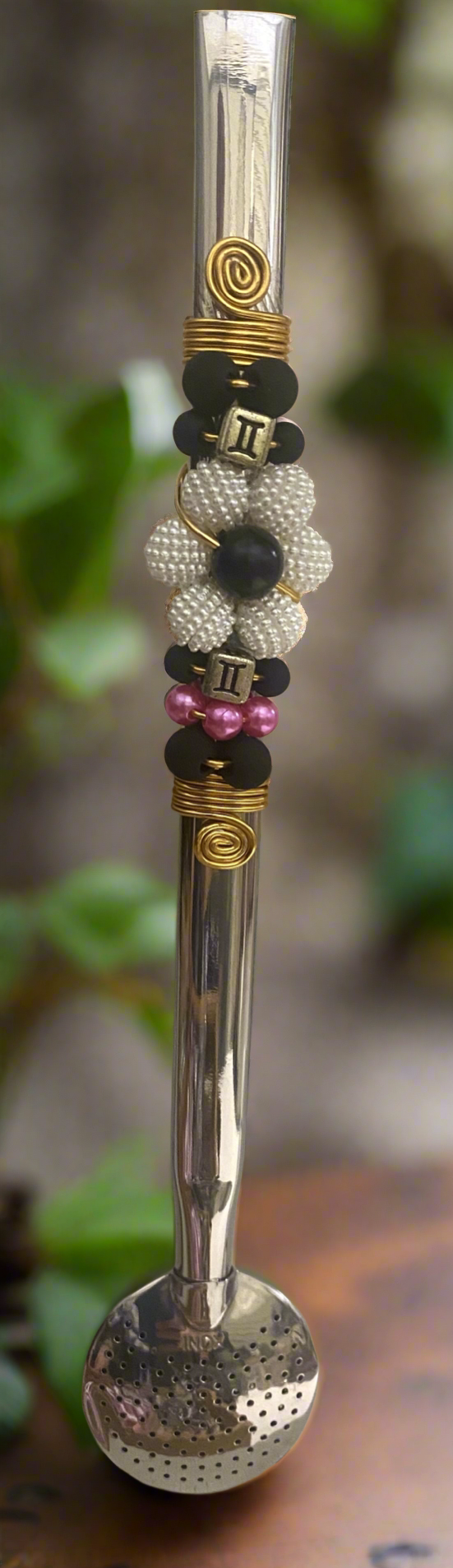 A close-up image of a stainless steel bombilla  with a floral pattern, featuring various colored beads and a yellow gold-tone band wiht a forest blurred background on a wood table. 