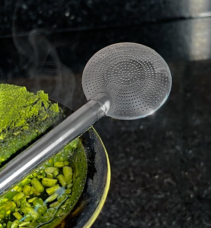 A stainless steel bombilla with 1000 holes used for siphoning yerba mate, shown with steam rising from the yerba and the tool partially submerged.