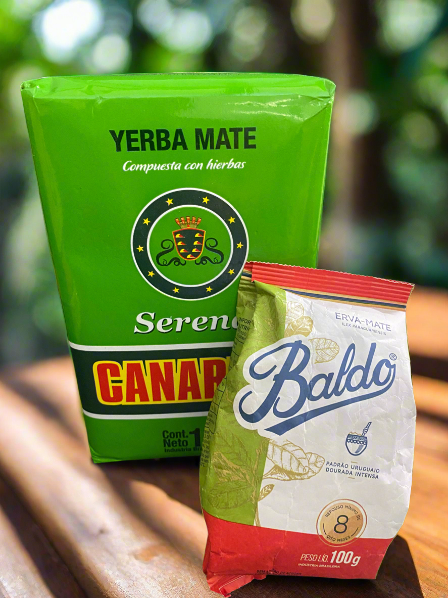 Two packages of yerba mate. One labeled 'Canarias Serena' in green and the other 'Baldo' in white and red, both on a wooden surface with foliage in the background.