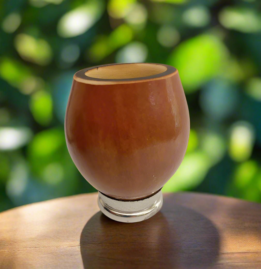 Yerba mate gourd with a metal base at bottom with a blurred leaf background on a wooden table.