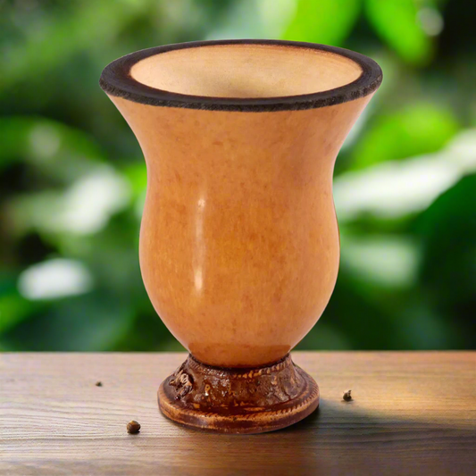 A traditional handmade gourd cuia, with a beige tone and decorative patterns at the base, displayed on a wooden surface with a blurred green background.