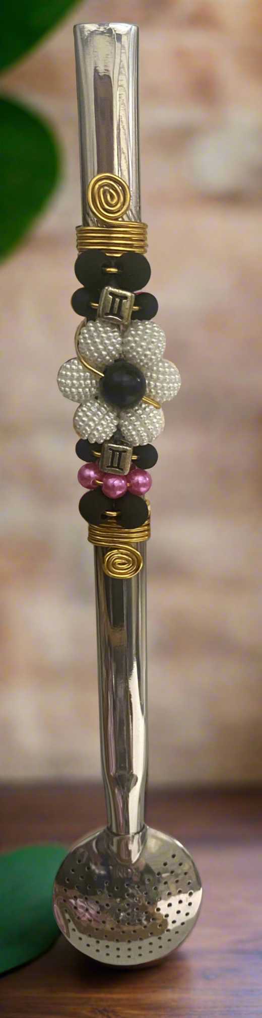 A close-up image of a stainless steel bombilla  with a floral pattern, featuring various colored beads and a yellow gold-tone band.