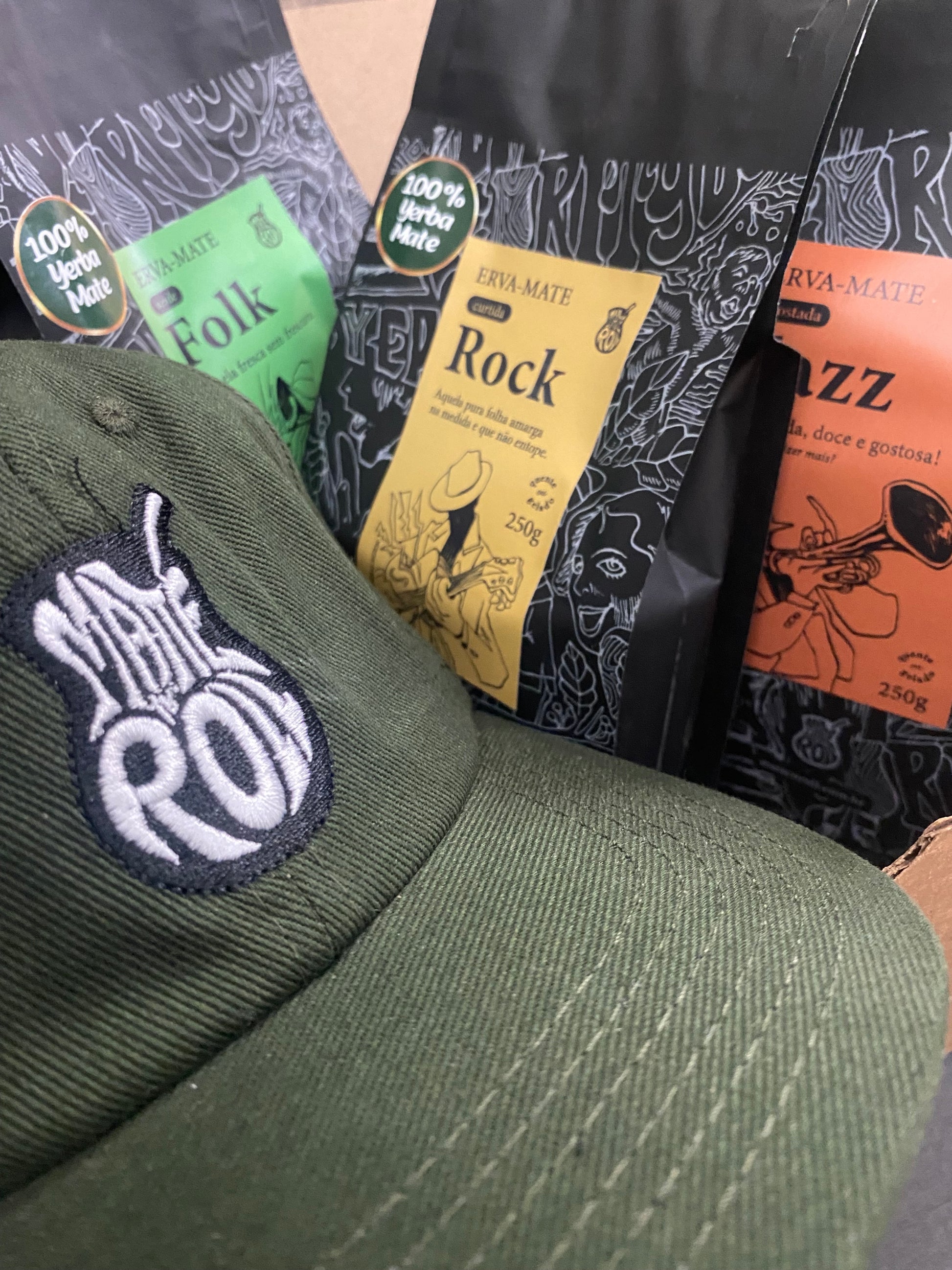 An assortment of Yerba Mate packages with different flavors, including 'ROCK', 'FOLK', and 'JAZZ', alongside a green hat with a logo.
