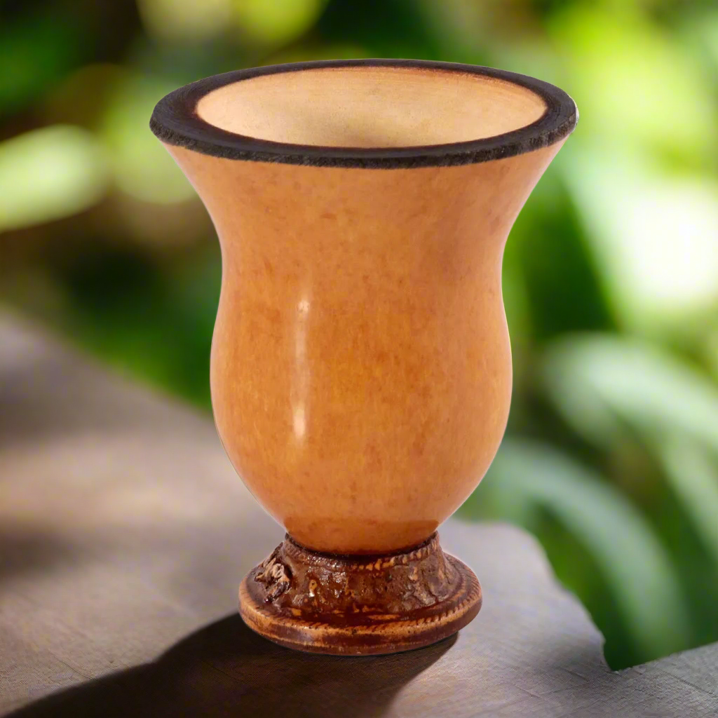 A handcrafted traditional gourd cup with a brownish color and patterned base, filled with a beige substance, set against a blurred green background.