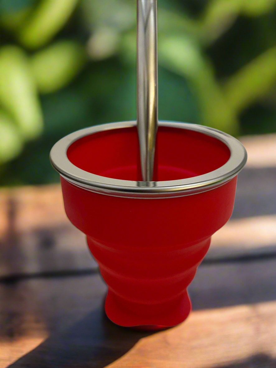 A red silicone collapsible mate with a silver-colored top and straw, placed on a wooden surface with foliage in the background.