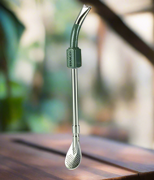 A green and silver metal bombilla, or straw, for yerba mate with a spring mechanism, displayed on a wooden surface with foliage in the background.