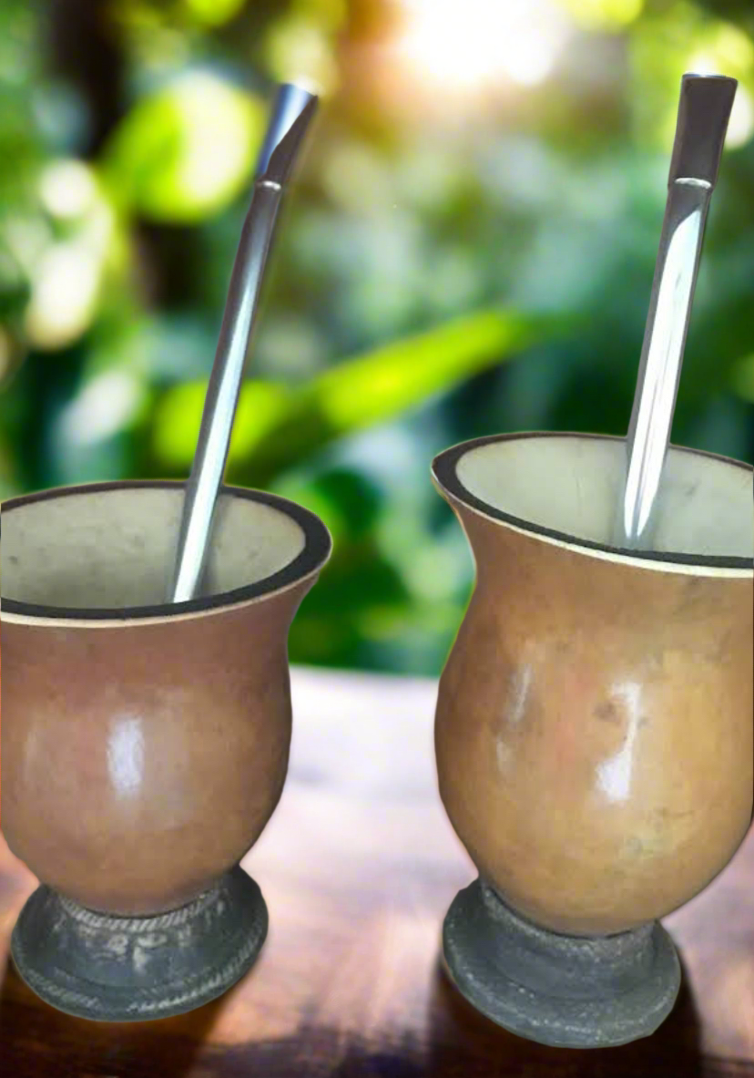 Two traditional gourd cups made from porongo, with a matte brown finish, displayed on a table with a blurred green background. One cup has a spoon in it, and the other is empty.